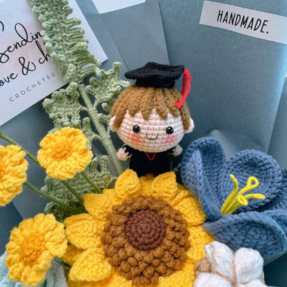 Handmade crochet figurine of a person in a graduation cap surrounded by crocheted flowers on a textured surface.