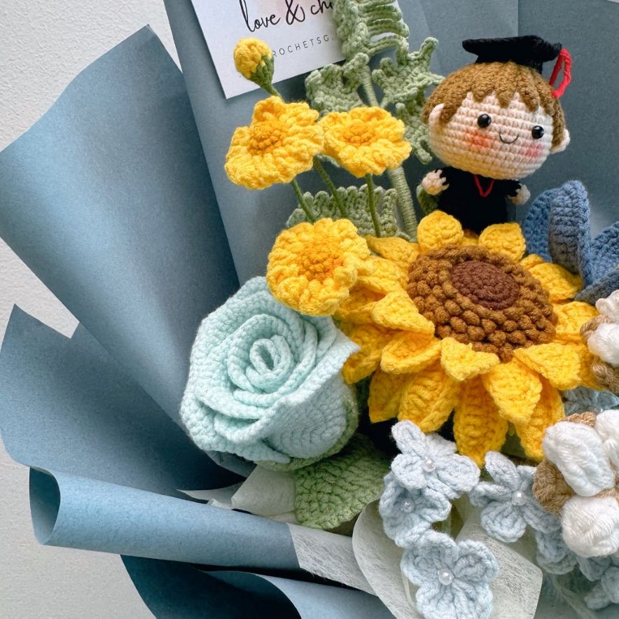 Crocheted bouquet with yellow sunflower, green leaves, and a small figurine in a graduation cap.