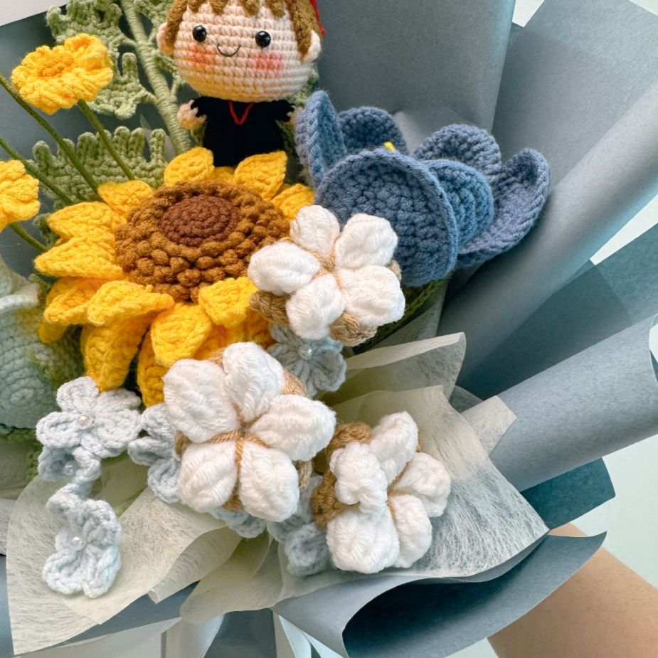 Crocheted sunflower, white flowers, and blue leaves with a small figurine on top.