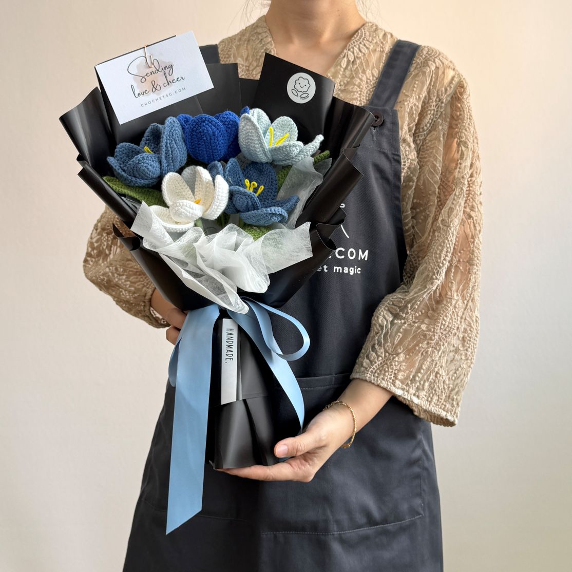 Person holding a bouquet of knitted flowers with a black card in a navy apron.