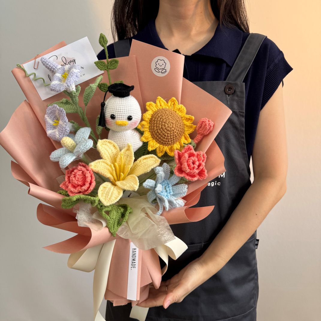 Person holding a bouquet of knitted flowers and a small plush toy against a neutral background
