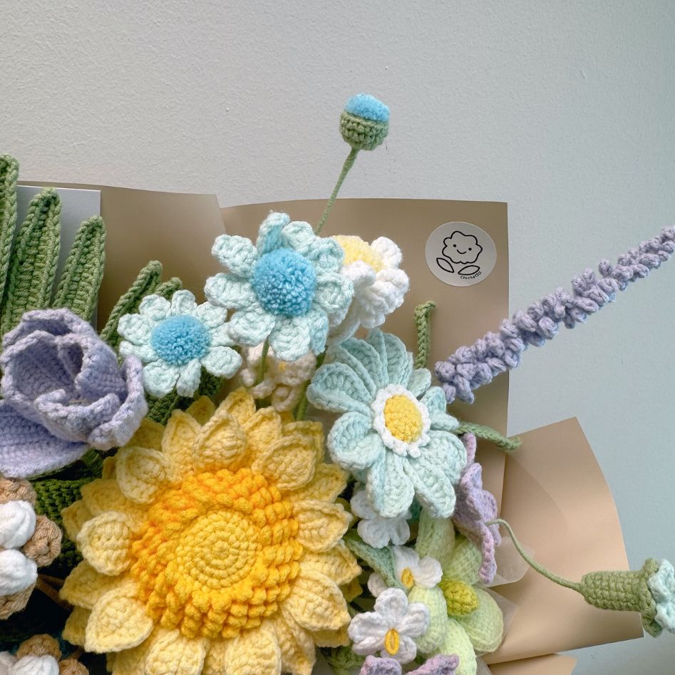 Crocheted flower bouquet with a yellow sunflower, blue and white flowers, and lavender on brown paper against a light gray background.
