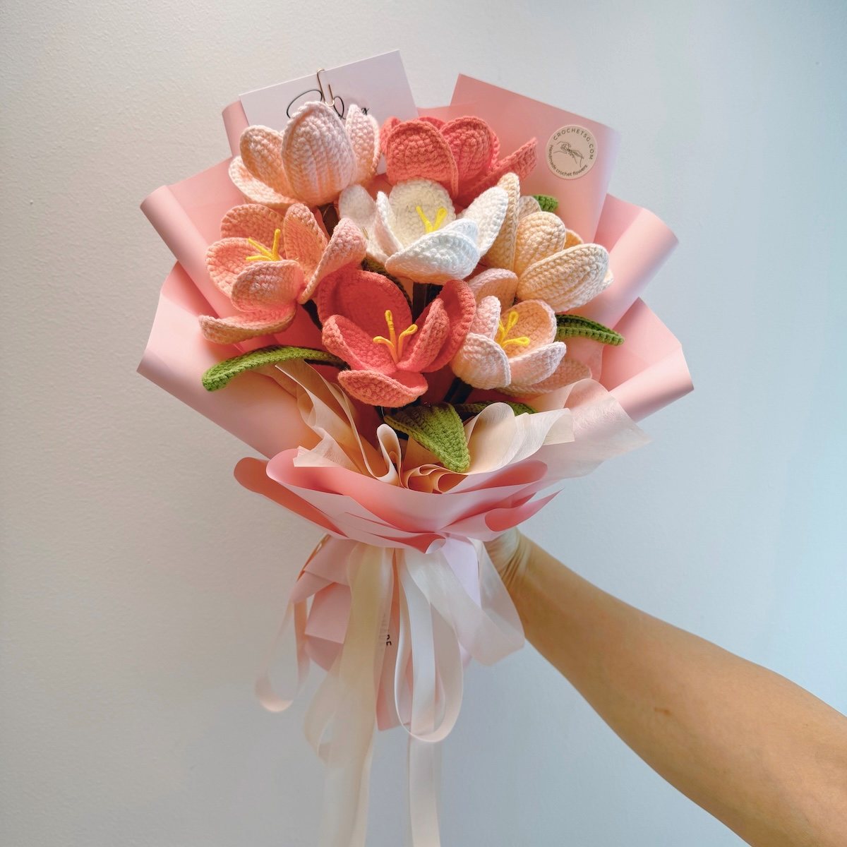 Bouquet of knitted flowers in pink, white, and green held by a hand against a light background